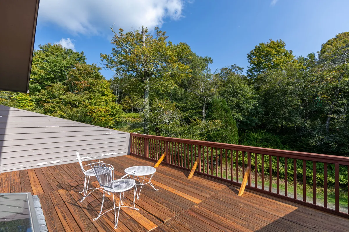 A wooden deck with white patio furniture overlooks lush greenery, inviting guests to relax and soak in the mountain environment. The setting is tranquil, with hints of open meadows and the surrounding forest just beyond.