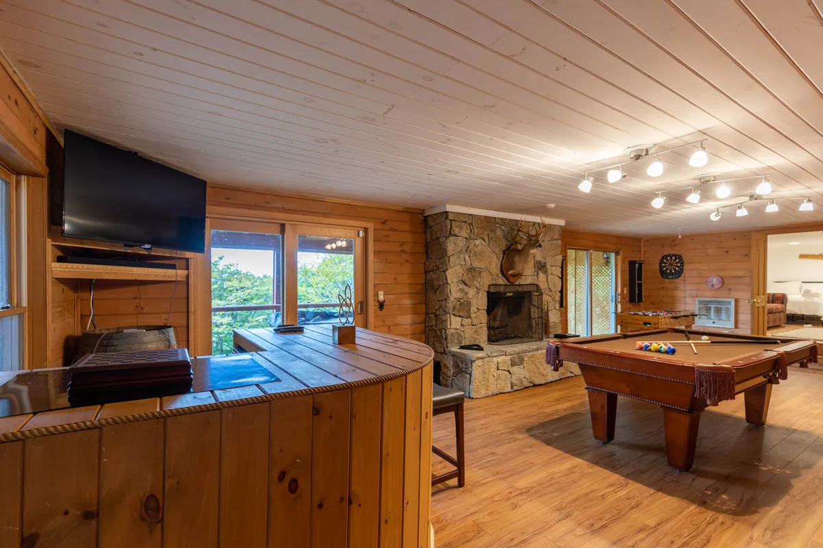 A spacious interior featuring a pool table at the center, a cozy bar area to the side, a stone fireplace along the back wall, and large windows letting in natural light. The furniture is arranged to facilitate both games and relaxed socializing, and the overall atmosphere is warm and inviting.