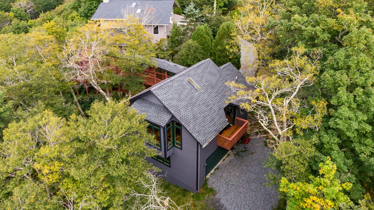 This image presents a broad exterior view of the property, highlighting the modern architectural style of the main house. The scene is set amid lush greenery, with a gravel path leading up to the entrance, and hints at the seamless blend of the home with its natural surroundings. Wide windows and the expansive setting suggest ample space and privacy for larger groups.
