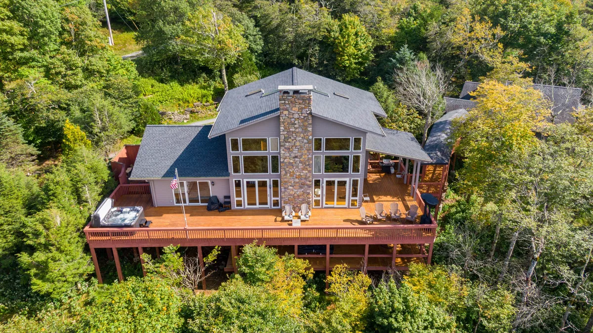 This exterior shot focuses on the side of the main house with its large windows, a stone chimney, and an inviting deck area framed by surrounding trees. The deck is ideal for gatherings, connecting the indoor living space with the outdoors, and the large windows suggest bright, communal interiors.