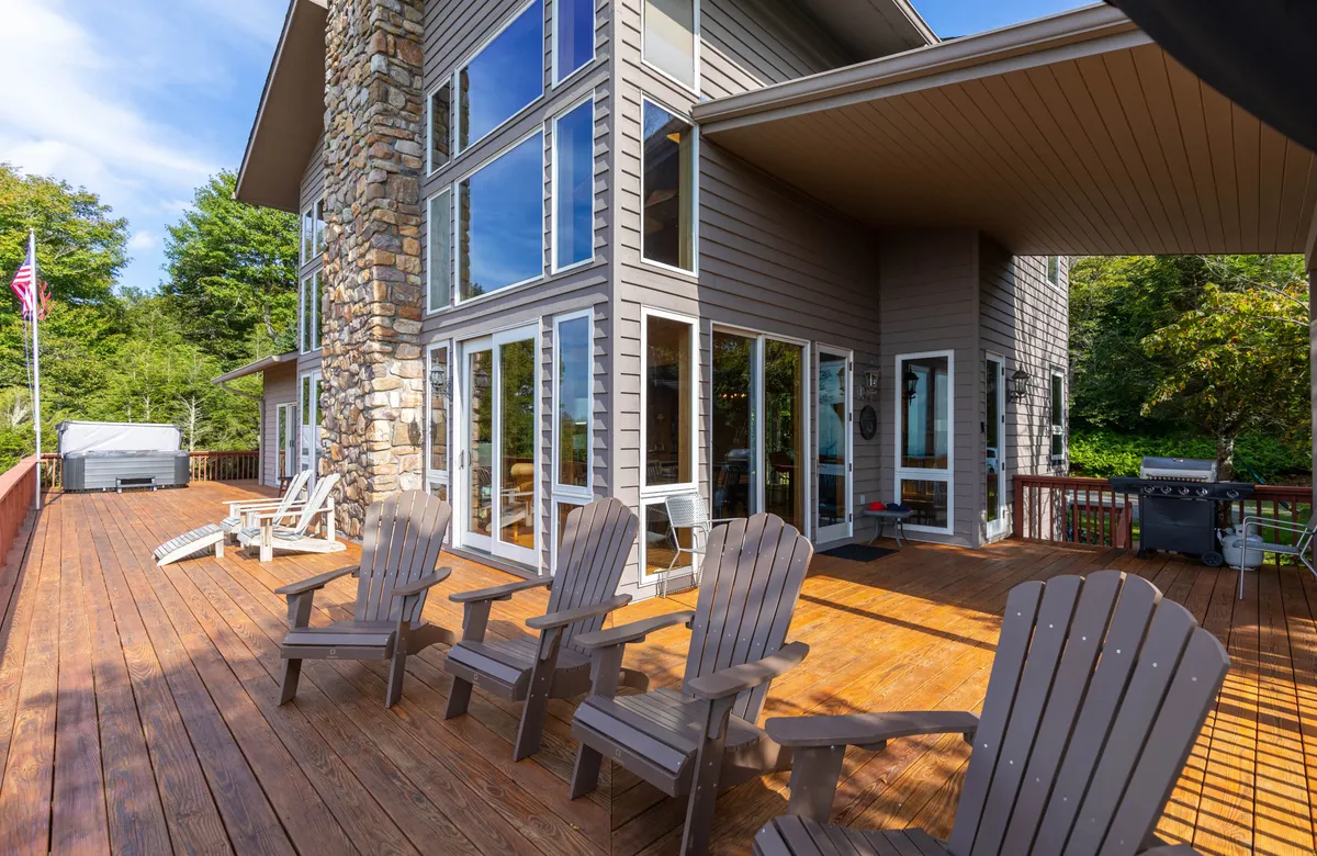 The exterior of the home features large windows and a spacious wooden deck furnished with adirondack chairs. The surrounding trees and stone chimney add rustic charm, while the deck invites social gatherings and outdoor meals. Sunsets and natural beauty are effortlessly accessible from this vantage point.