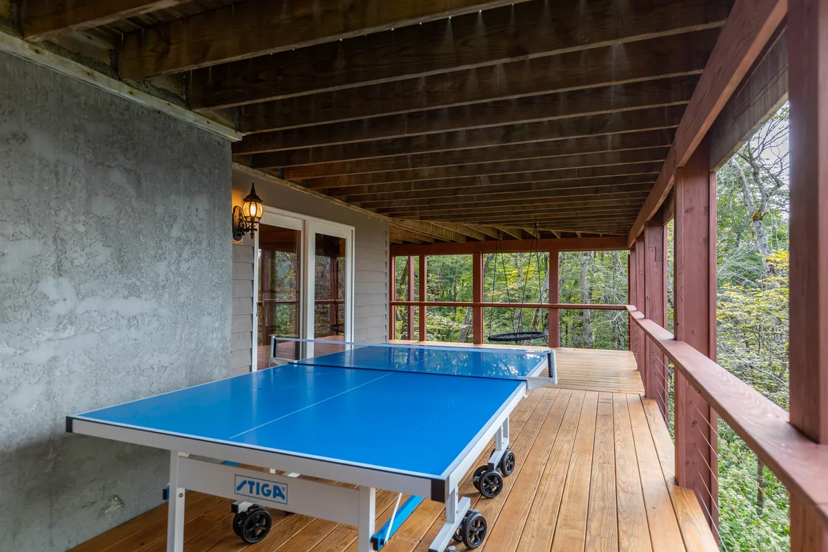 A lively exterior scene showcasing a ping pong table on a spacious wooden deck, surrounded by trees. The glass doors open to the interior, blending indoor and outdoor experiences and suggesting social gatherings or family fun against the backdrop of the mountains.