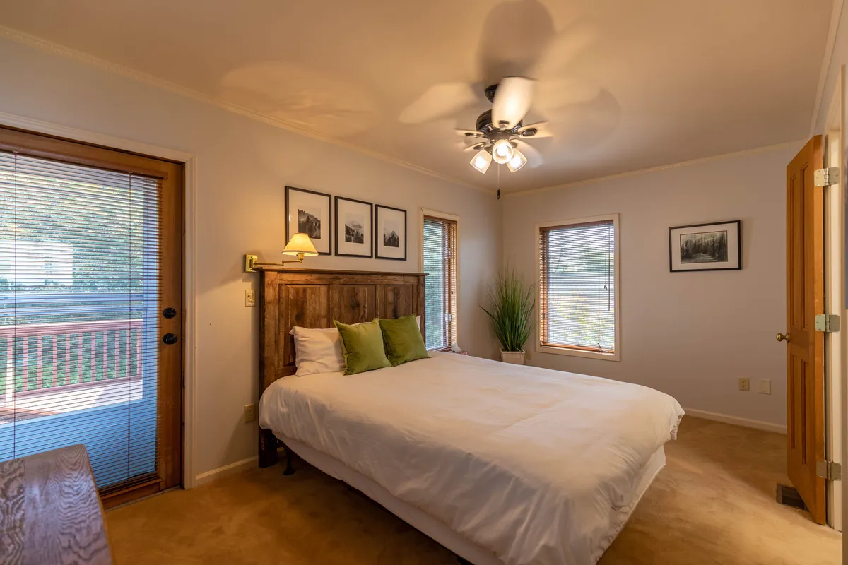 A cozy bedroom with a wooden headboard, framed pictures, and large windows that provide views of nature. The room is bright and inviting, offering an intimate retreat within the cottage.