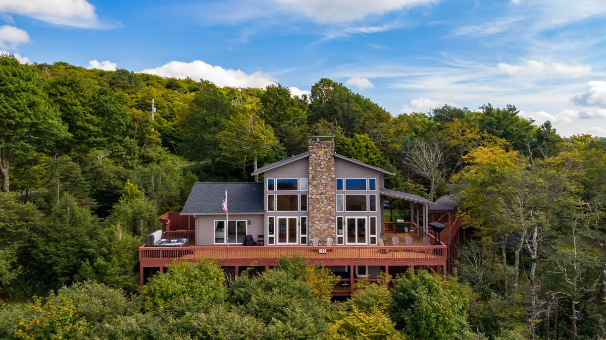 The home’s exterior features large windows and a stone chimney, opening out to a spacious wooden deck surrounded by lush forest. The composition includes outdoor furniture, inviting guests to relax and take in sunset views or host intimate gatherings while immersed in nature.