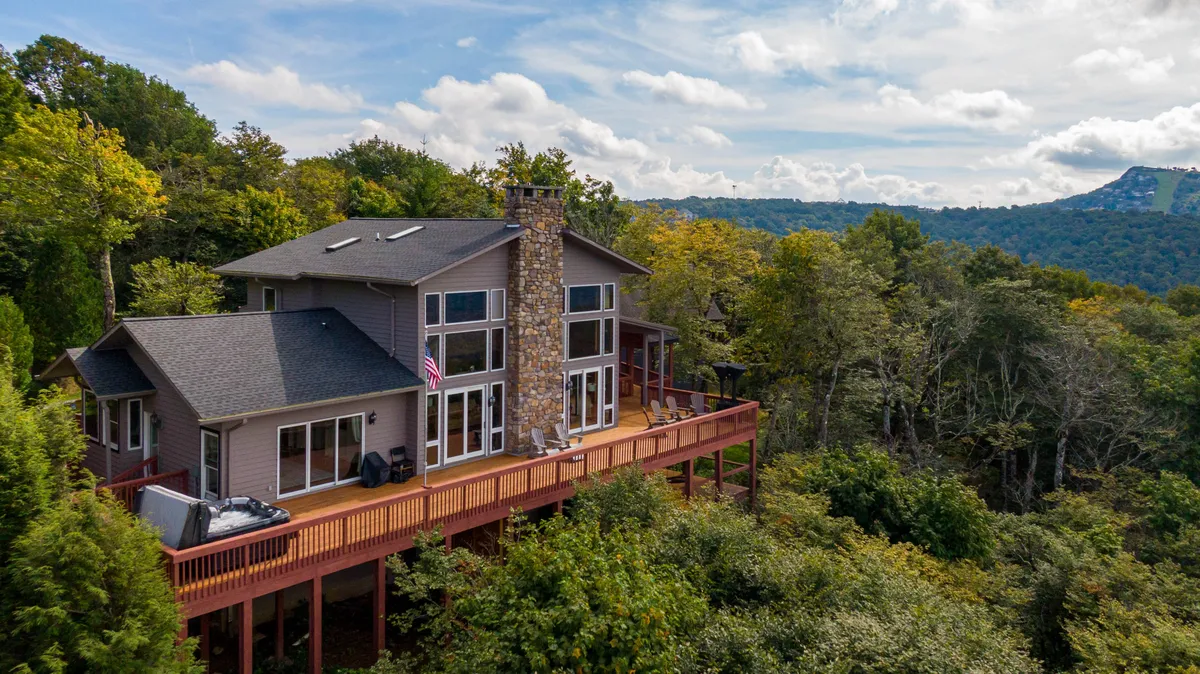 The image showcases the stunning exterior of Skytop Chateau, featuring a spacious deck with outdoor dining options, a striking stone chimney, and expansive windows that capture panoramic mountain views. The home is nestled among lush trees, set atop 5 private acres, with the deck offering a perfect vantage point to take in vibrant sunsets or enjoy morning coffee immersed in nature.