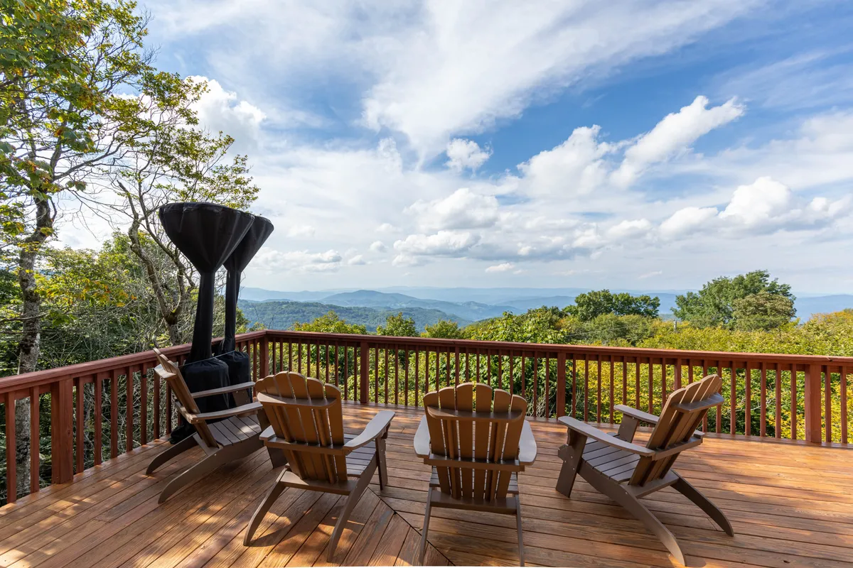 A serene wooden deck furnished with classic adirondack chairs faces a sweeping mountain vista beneath a cloudy sky. The setting is inviting for small groups or individuals to relax, socialize, or simply take in the natural beauty. The edge of the deck blends seamlessly with the surrounding landscape, suggesting easy access to outdoor activities and nature.