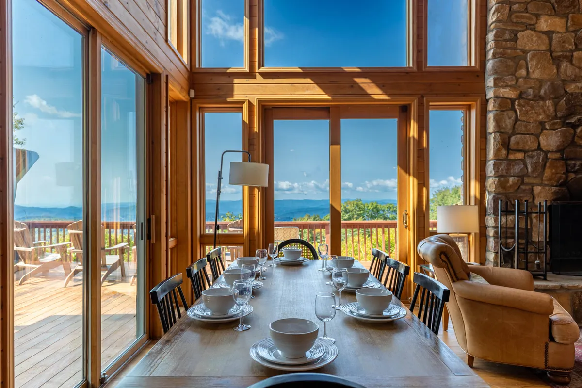 A large dining table set for a meal stands next to expansive windows that showcase sweeping mountain vistas. The stone fireplace adds a touch of rustic elegance, while the inviting atmosphere is perfect for shared dinners and celebrations with loved ones.