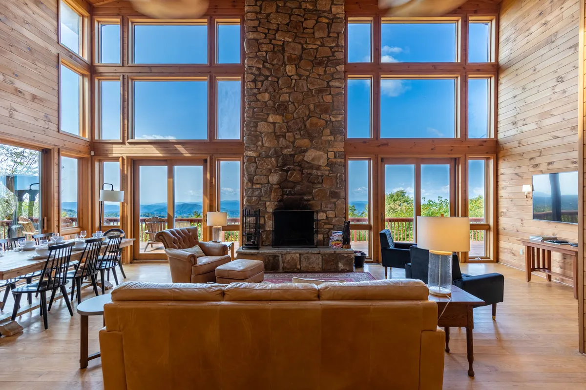 This image features a warm and welcoming great room with an impressive large stone fireplace as the focal point. Expansive windows frame breathtaking mountain views, while the comfortable seating area and adjacent dining table invite guests to gather and relax. The scene conveys togetherness, warmth, and the joy of shared moments.