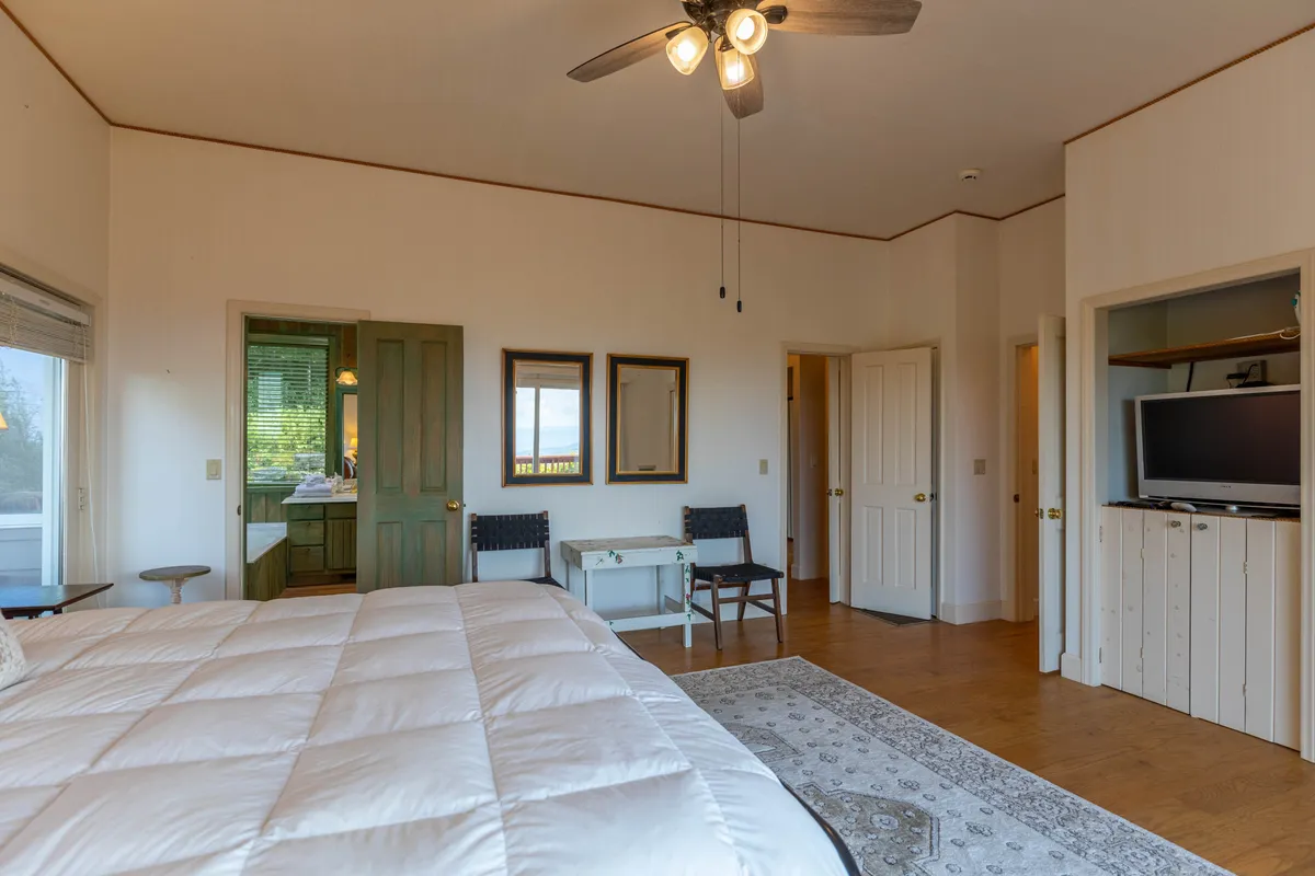A spacious king-sized bed with crisp white bedding sits against a backdrop of warm wooden accents. The room is filled with natural light, and a green door leads directly to an ensuite bathroom. Two framed mirrors and a small table with chairs add to the suite-like ambiance, conveying comfort and easy access to amenities.
