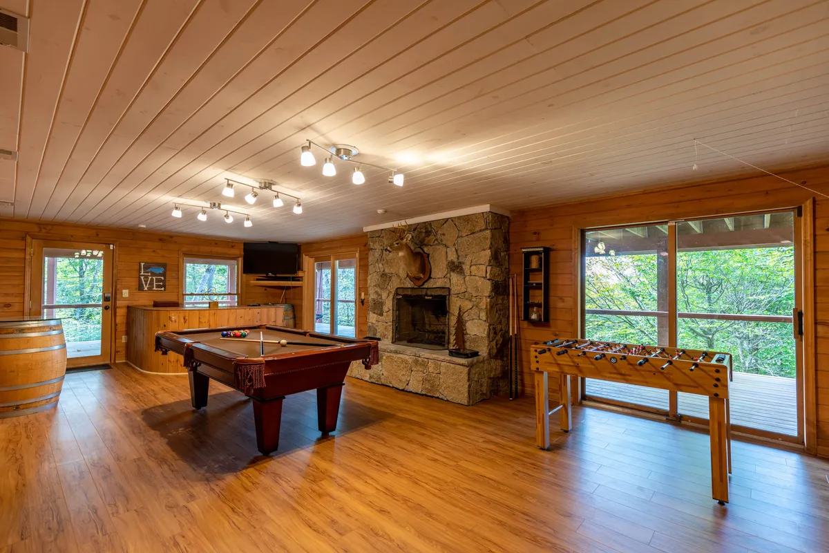 A vibrant entertainment room with a pool table, foosball table, wood-burning fireplace, and large windows offering views of the outdoors. The layout is casual and conducive to group activities, with plenty of space for movement and gathering.