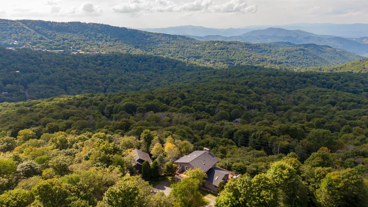 A sweeping aerial view showcases the spacious Skytop Chateau perched atop a densely forested mountain, with layers of blue-hued peaks stretching into the distance. The image captures both the grand scale of the home and its peaceful isolation within acres of private woods, emphasizing the dramatic elevation and scenic mountain setting.
